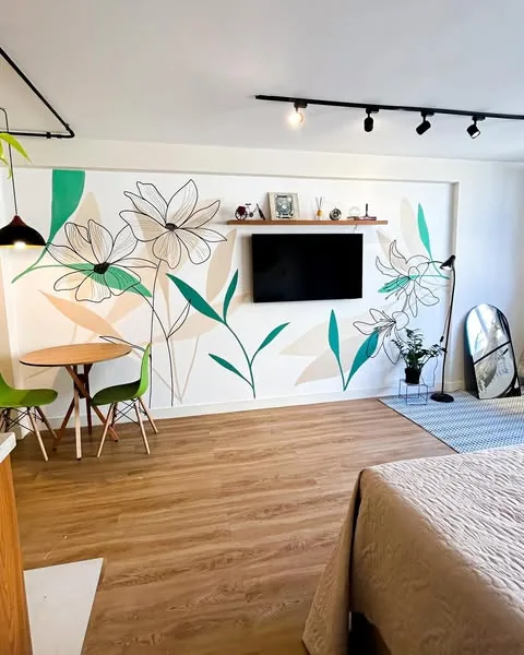 Mural floral em contornos pretos com folhas verdes e formas bege na parede de uma sala com TV
